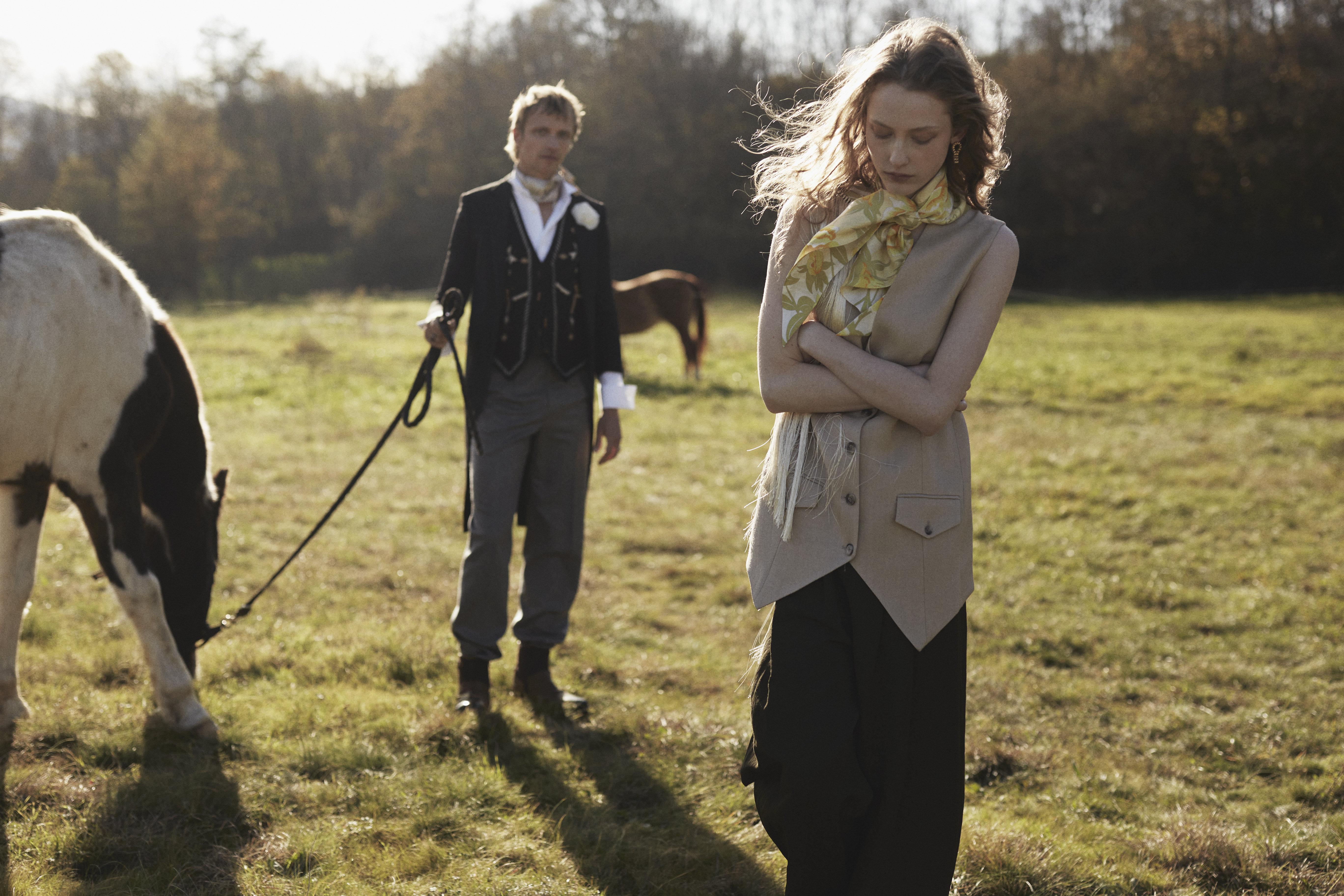 A fashionable couple poses with horses in a sunlit field, with the woman in a tan vest and black pants and the man in a tuxedo.
