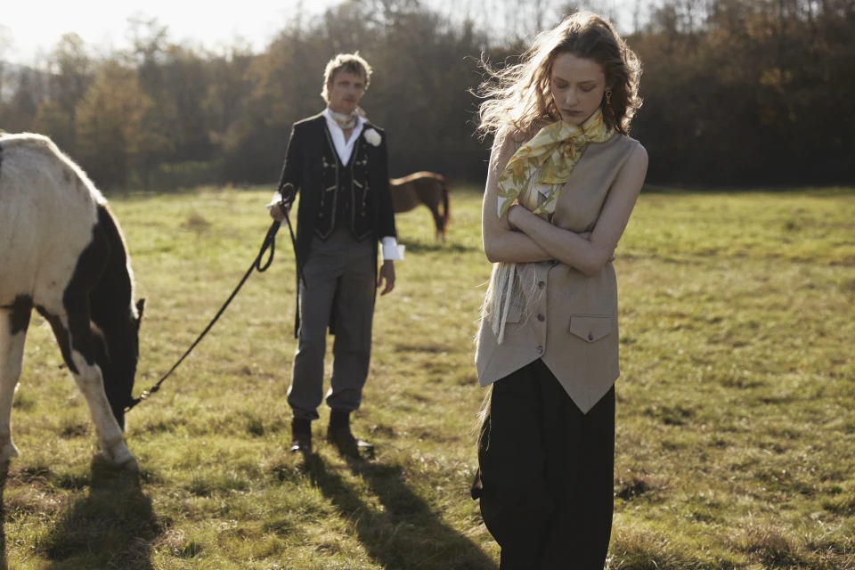 A fashionable couple poses with horses in a sunlit field, with the woman in a tan vest and black pants and the man in a tuxedo.