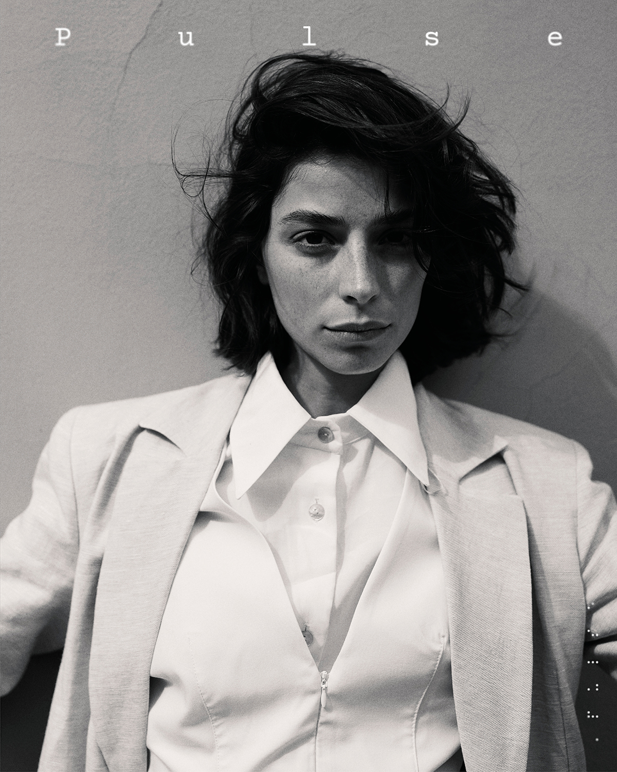 Headshot portrait of a brunette woman with a short, textured hairstyle, wearing an open white blouse and jacket against a neutral wall.