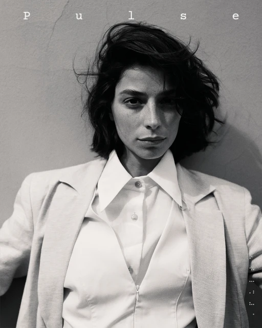 Headshot portrait of a brunette woman with a short, textured hairstyle, wearing an open white blouse and jacket against a neutral wall.