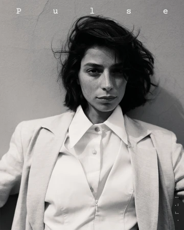 Headshot portrait of a brunette woman with a short, textured hairstyle, wearing an open white blouse and jacket against a neutral wall.