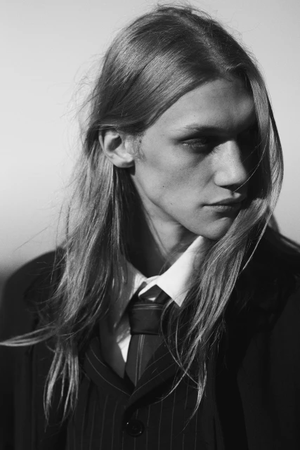 Monochrome portrait of a young adult male with long hair wearing a pinstripe suit, white collared shirt, and tie, posing in a photo studio.