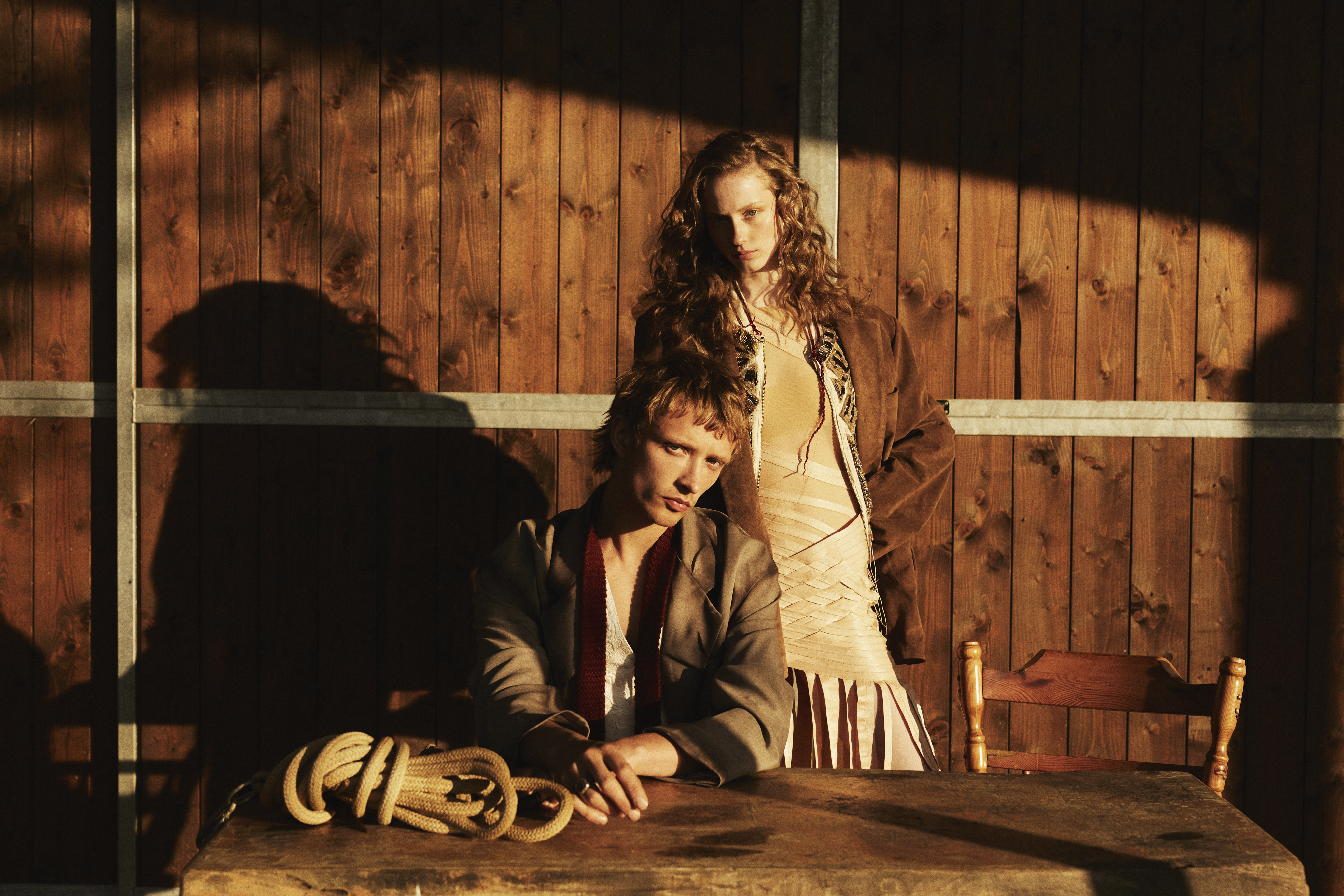 Two fashion models pose together in front of a rustic wood background with harsh shadows and warm tones for an editorial shoot.