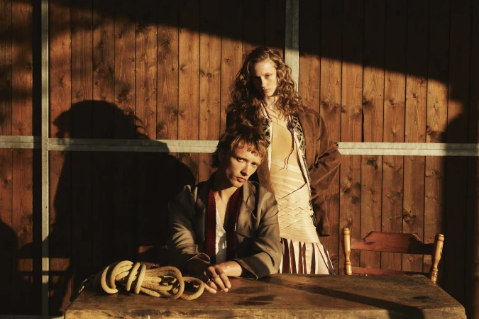 Two fashion models pose together in front of a rustic wood background with harsh shadows and warm tones for an editorial shoot.