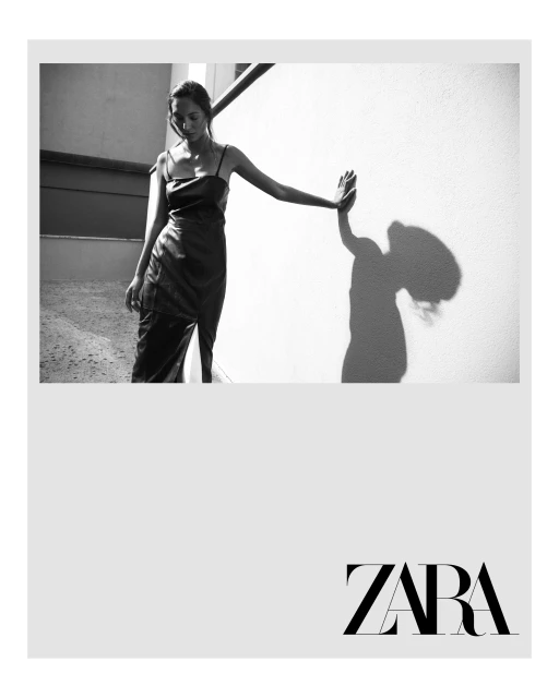 A solitary adult female in a sleek dress poses with her hand against a textured wall, casting a shadow in a black and white fashion shot.