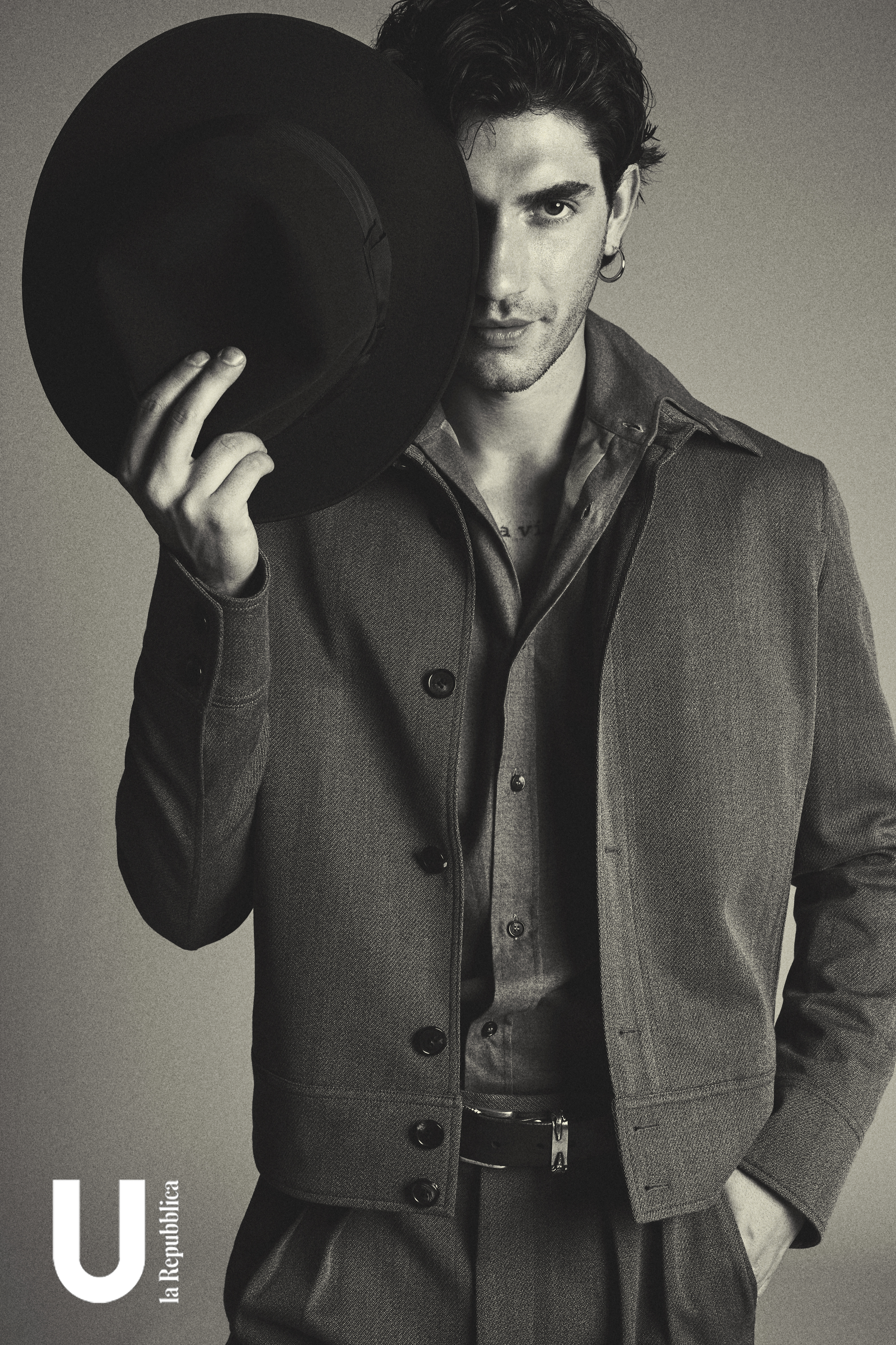 Portrait of a male fashion model with dark hair partially obscured by a black hat, wearing a brown jacket and shirt, against a grey background.