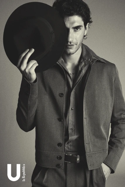 Portrait of a male fashion model with dark hair partially obscured by a black hat, wearing a brown jacket and shirt, against a grey background.