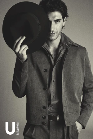 Portrait of a male fashion model with dark hair partially obscured by a black hat, wearing a brown jacket and shirt, against a grey background.