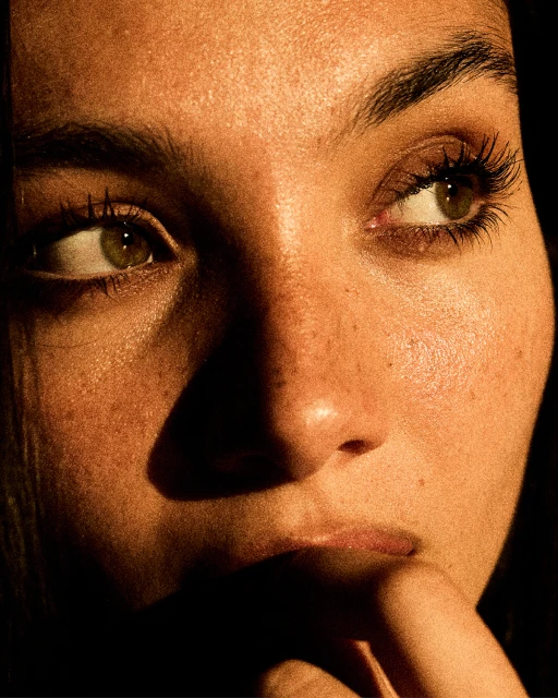 Close-up portrait of a young adult woman with green eyes, dark hair, and freckles, illuminated by warm, directional lighting.