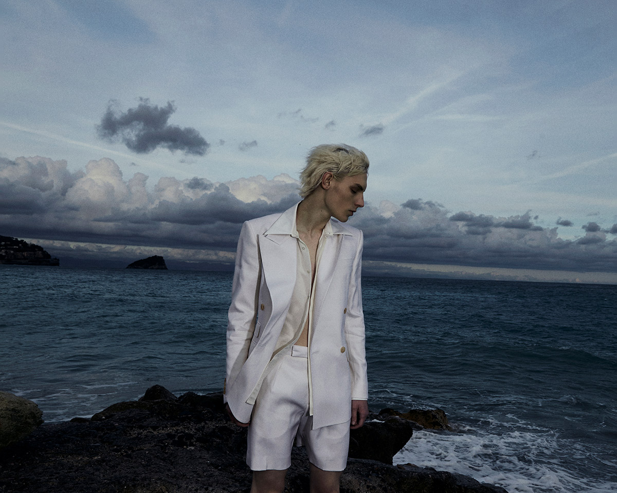 Fashionable male model with blond hair posing in a white suit jacket and shorts on a rocky shore with a stormy ocean backdrop.