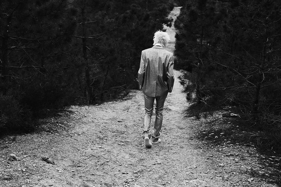 Back view of stylish adult in suit walking a gravel path through a dense forest in black and white. Fashion, nature, and solitude.