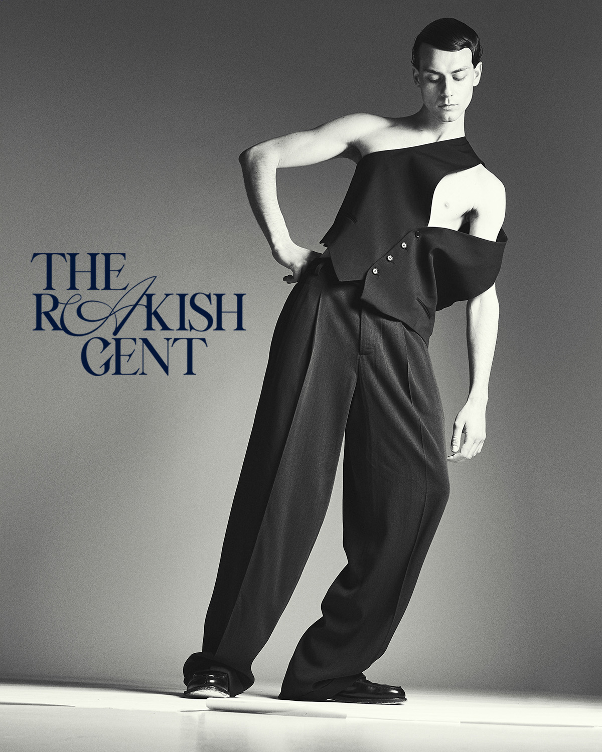 Fashionable male model in asymmetrical black vest and wide-leg trousers poses in a studio with modern style and gray background.
