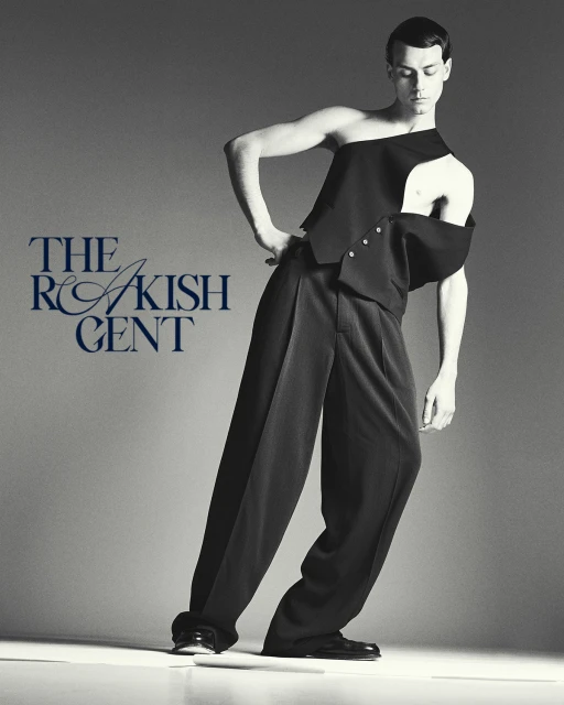 Fashionable male model in asymmetrical black vest and wide-leg trousers poses in a studio with modern style and gray background.