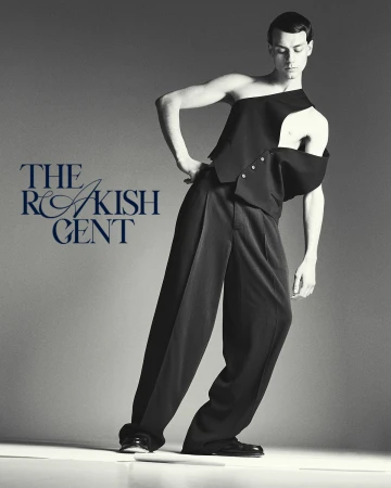 Fashionable male model in asymmetrical black vest and wide-leg trousers poses in a studio with modern style and gray background.