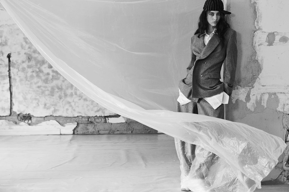 Black and white fashion shot of a woman in a suit and baseball cap, posing near a weathered wall with a translucent sheet blowing.
