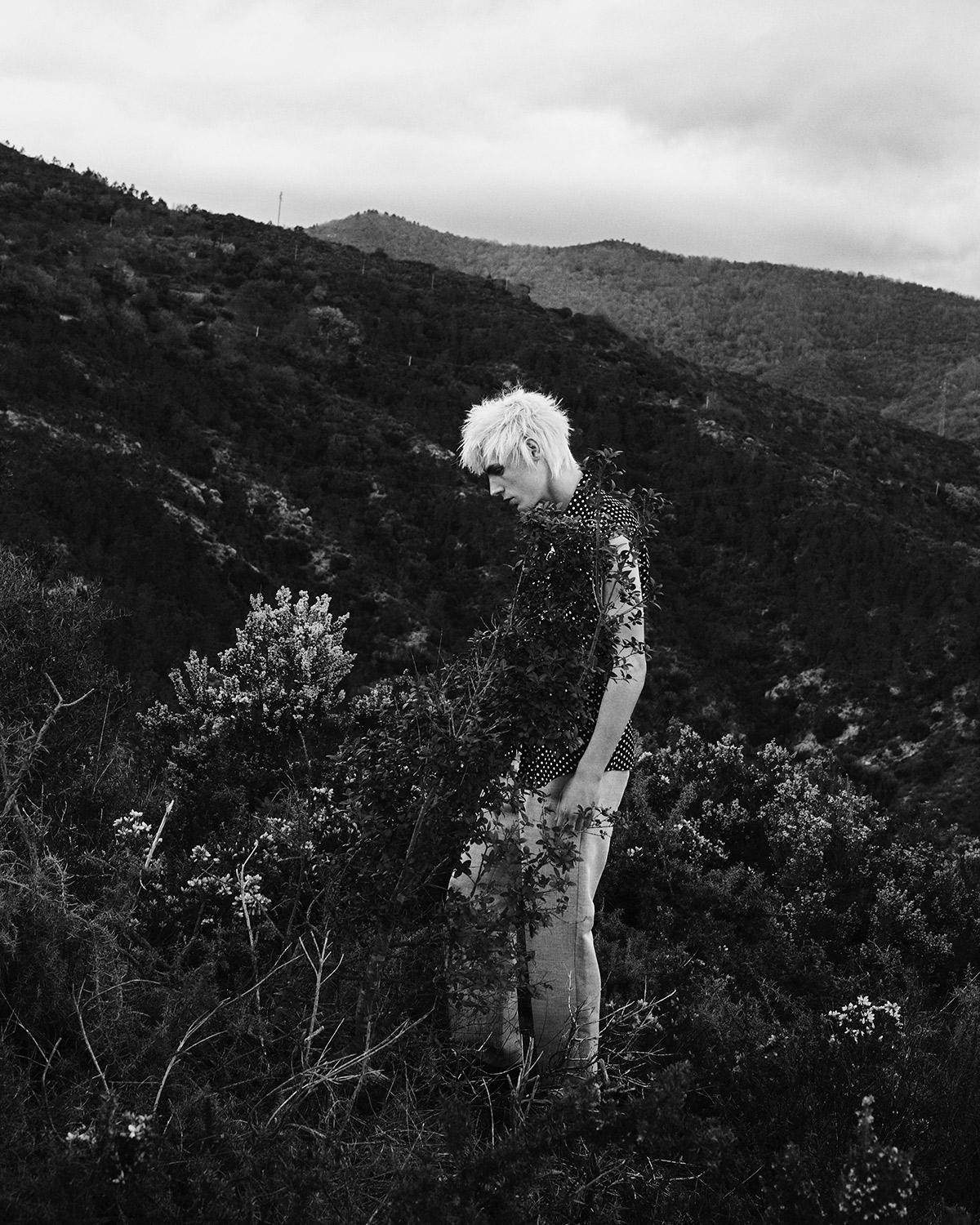 Monochrome portrait of a person with a light complexion, spiky hair, and polka-dot shirt standing amidst a grassy hillside under an overcast sky.