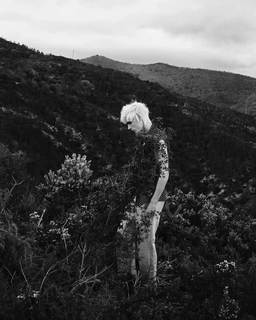 Monochrome portrait of a person with a light complexion, spiky hair, and polka-dot shirt standing amidst a grassy hillside under an overcast sky.