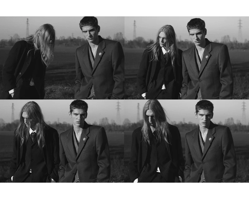 Black and white fashion shot of two young adult men in suits standing outdoors in a field, with powerlines in the background. 