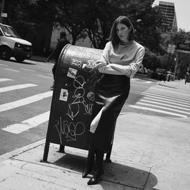 A woman in a skirt and top stands on a city sidewalk leaning against a public mailbox in a black and white street fashion image.