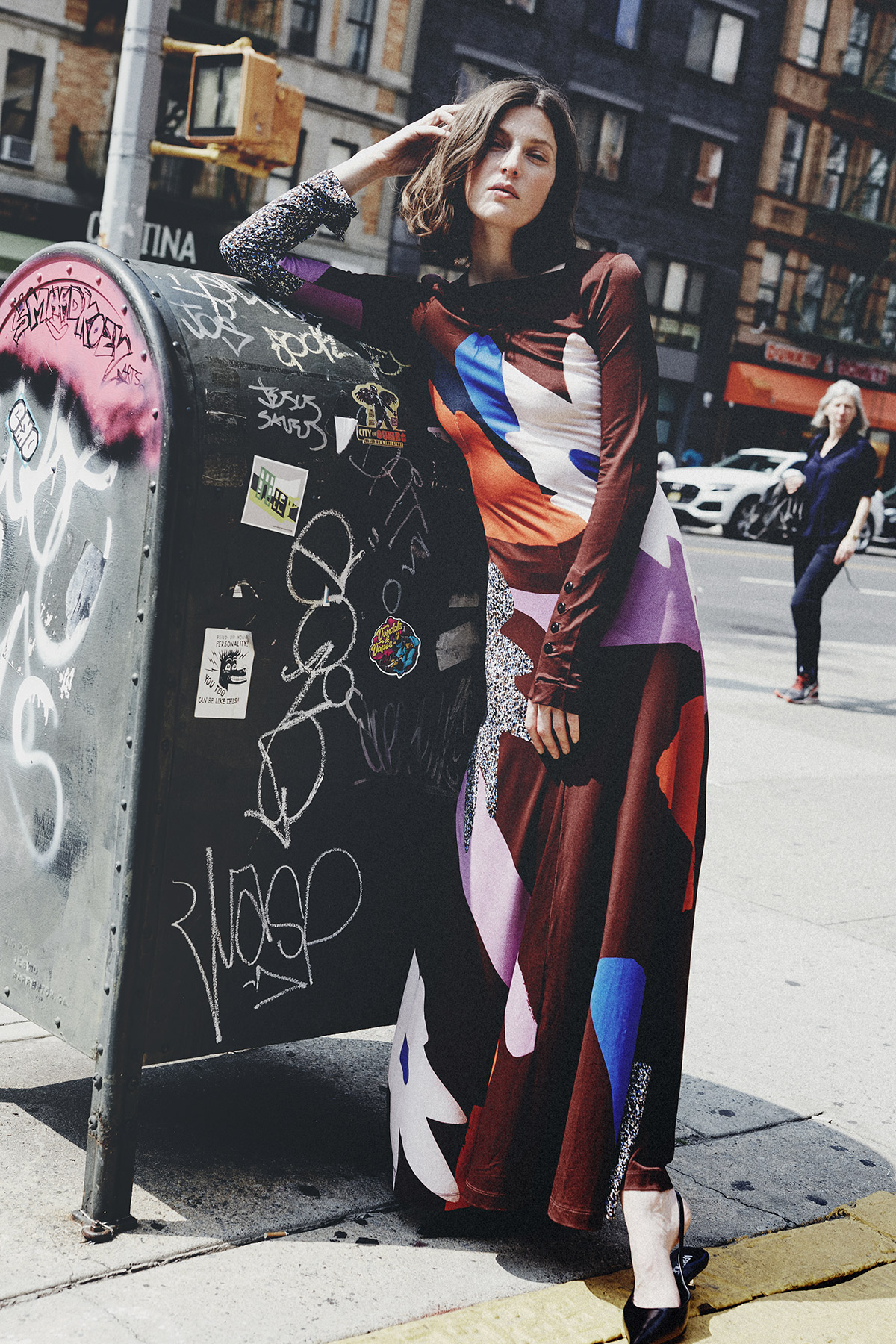 A woman in a long, patterned dress poses on a city sidewalk, leaning against a graffiti-covered mailbox, under a sunny sky.