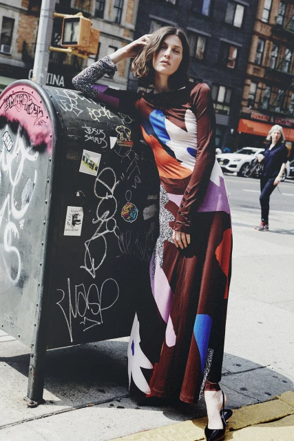 A woman in a long, patterned dress poses on a city sidewalk, leaning against a graffiti-covered mailbox, under a sunny sky.