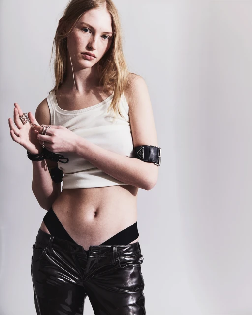 A fair-skinned young woman with long blonde hair poses in a photo studio, wearing a tank top, shiny pants, multiple rings, and a black arm cuff.