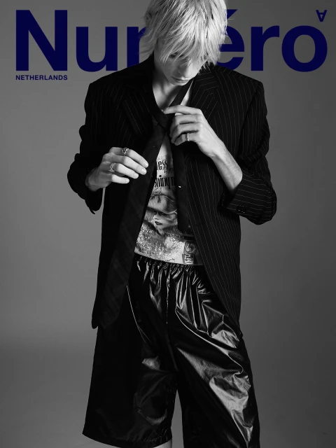 Blonde male model in a pinstripe blazer and black shorts poses for a fashion shot with the magazine Numero Netherlands logo in the background.
