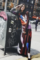 Woman in patterned long-sleeve dress leaning against a graffiti-covered mailbox on a city street