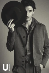 Man holding a black fedora hat partially covering his face while wearing a collared button-up shirt and jacket