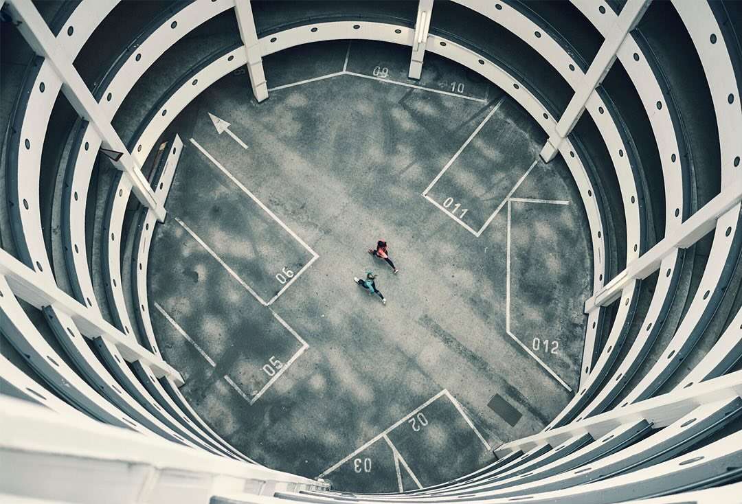 Overhead shot of two people in a concrete parking structure, walking toward each other, with white lines and architectural details.
