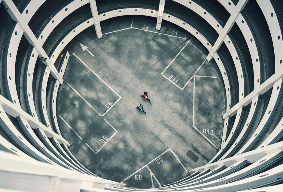 Overhead shot of two people in a concrete parking structure, walking toward each other, with white lines and architectural details.