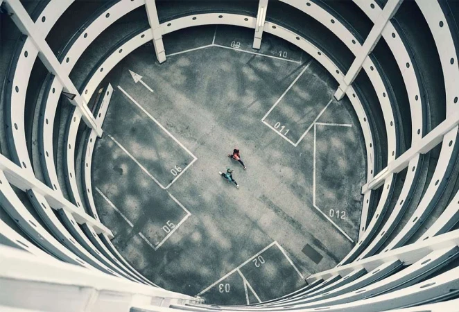 Overhead shot of two people in a concrete parking structure, walking toward each other, with white lines and architectural details.