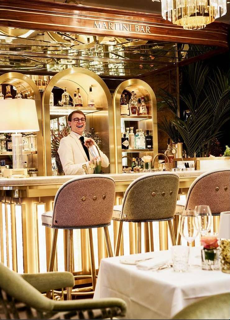 A bartender in a cream-colored jacket mixes drinks at a bar with golden accents, high chairs, and a chandelier in a warm indoor setting.