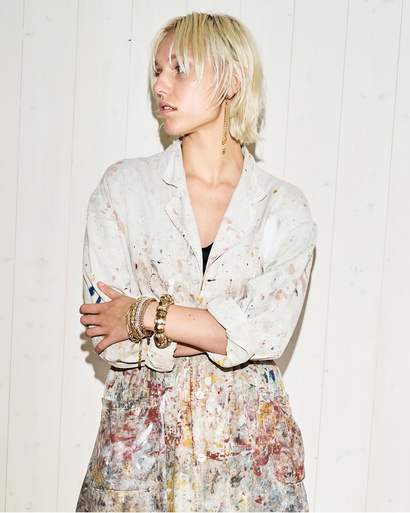 Blonde woman with painter's smock in a studio setting, accessorized with gold jewelry.  Fashion and beauty shot with neutral tones.