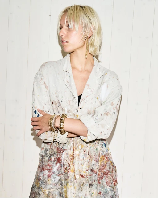 Blonde woman with painter's smock in a studio setting, accessorized with gold jewelry.  Fashion and beauty shot with neutral tones.