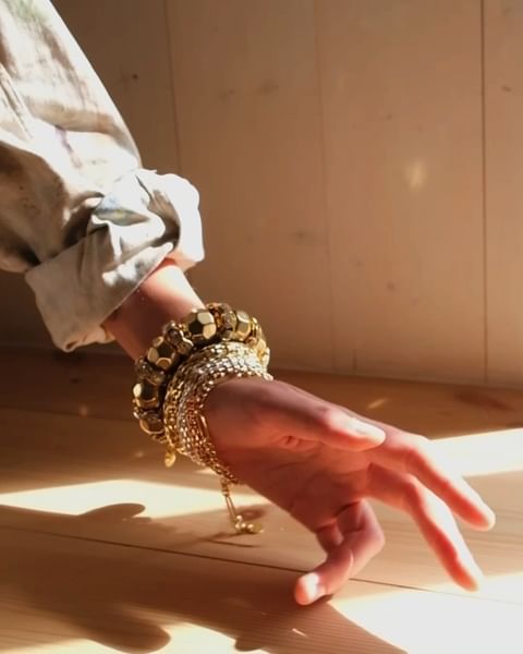 Close-up of a woman's hand adorned with multiple gold bracelets resting on a wooden surface in a naturally lit, warm environment.