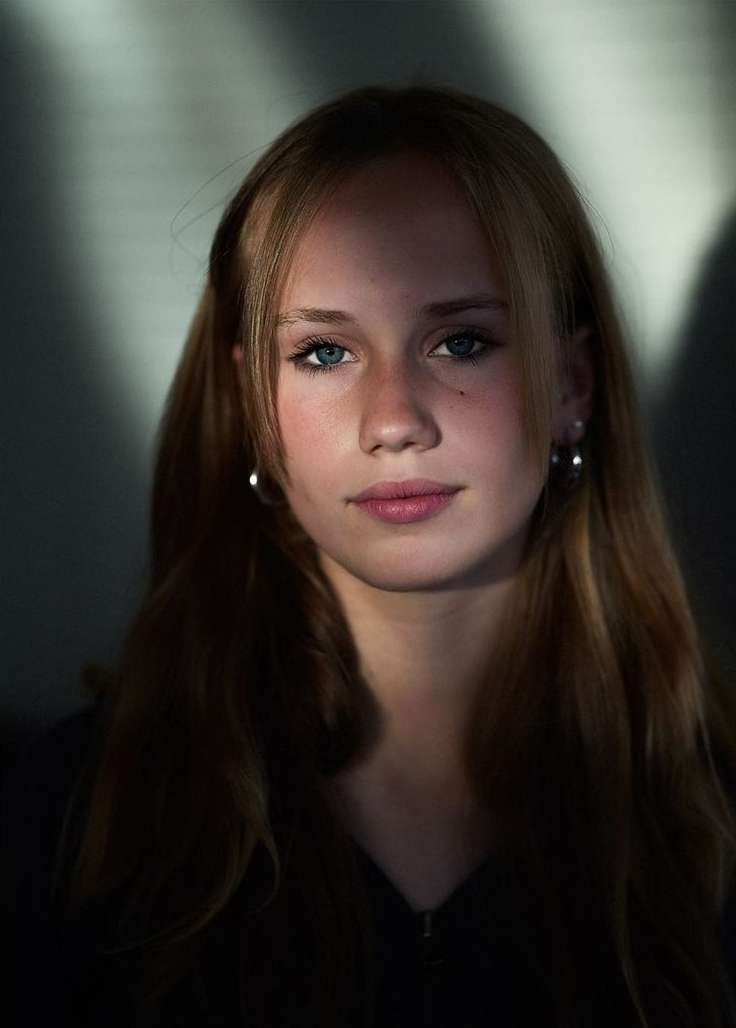 Portrait of a young woman with fair hair and blue eyes, featuring soft, diffused lighting and a simple dark top against a neutral background.