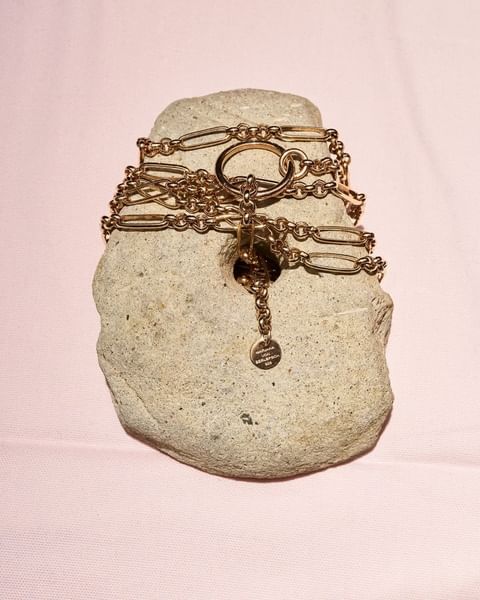 Close-up of a gold chain necklace draped on a textured gray rock against a soft pink background in a jewelry product shot.
