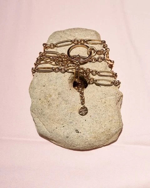 Close-up of a gold chain necklace draped on a textured gray rock against a soft pink background in a jewelry product shot.