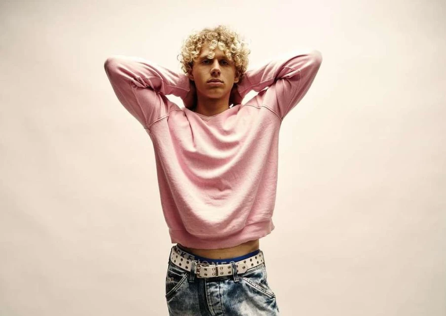 Young adult male with blonde curly hair, wearing a pink sweatshirt and jeans, poses against a plain background in a studio shot.