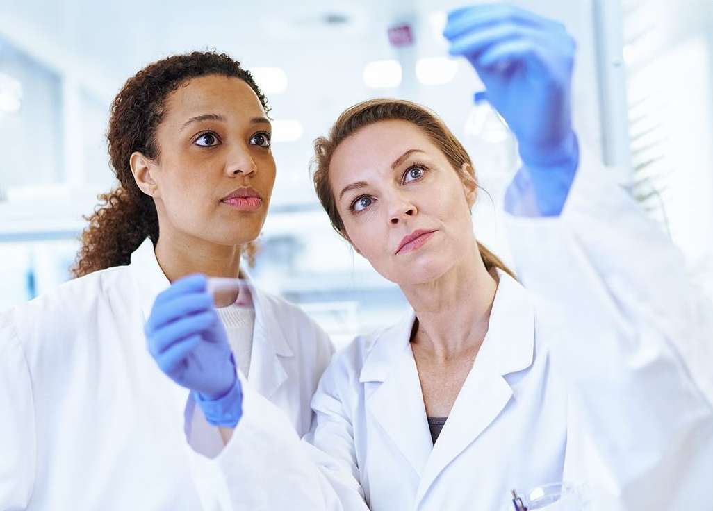 Two female scientists in lab coats and gloves examine a vial in a bright laboratory, with light reflecting off the glass.