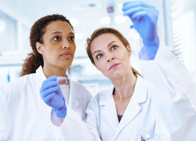 Two female scientists in lab coats and gloves examine a vial in a bright laboratory, with light reflecting off the glass.