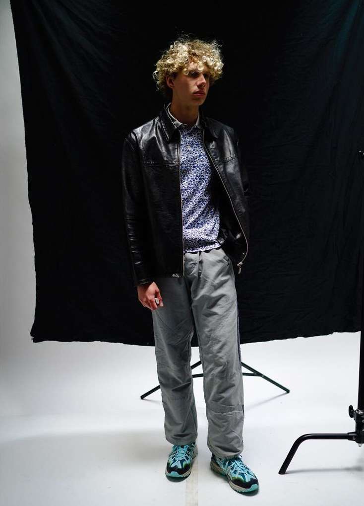 Full-length shot of a young male adult with curly hair in a leather jacket and patterned shirt, standing in front of black backdrop.