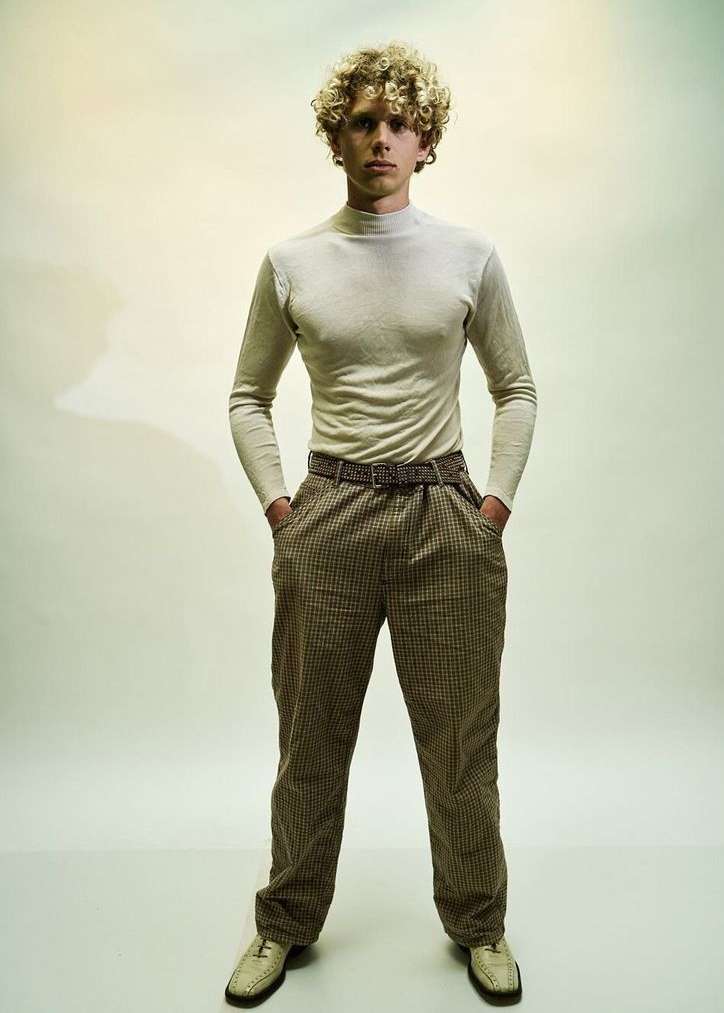 Full-length portrait of a young adult male with curly blonde hair, wearing a turtleneck, patterned pants, and leather loafers in a studio.
