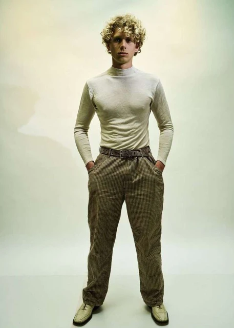 Full-length portrait of a young adult male with curly blonde hair, wearing a turtleneck, patterned pants, and leather loafers in a studio.