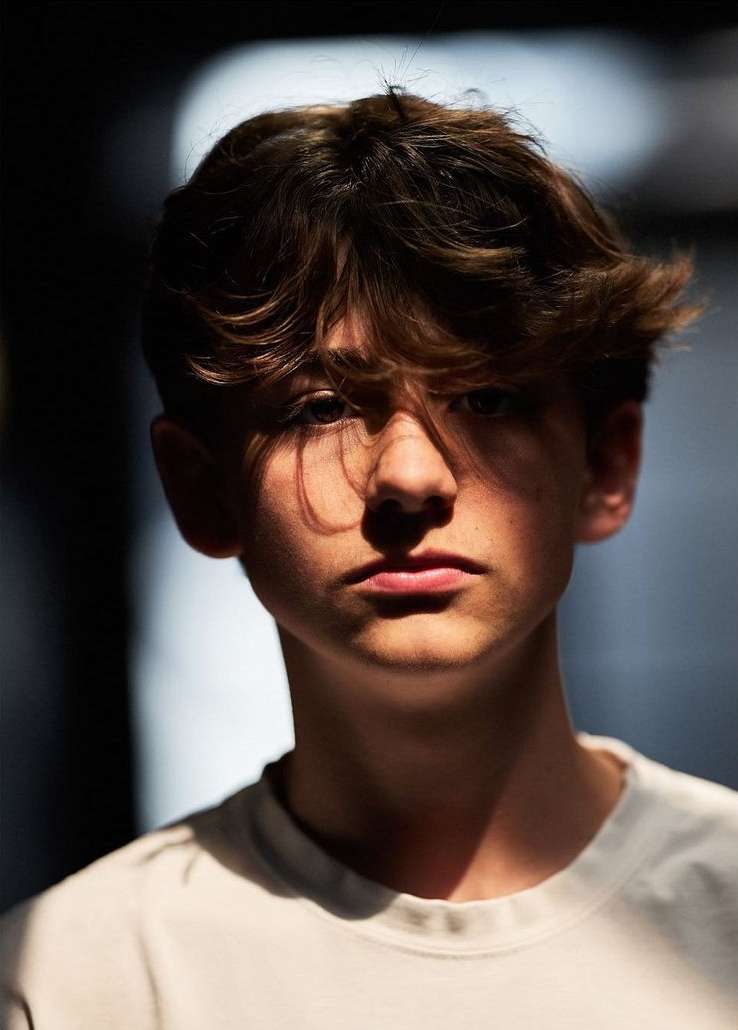 Portrait of a teenage boy with light brown hair and a white t-shirt. Shadows create contrast on his face in an indoor setting.