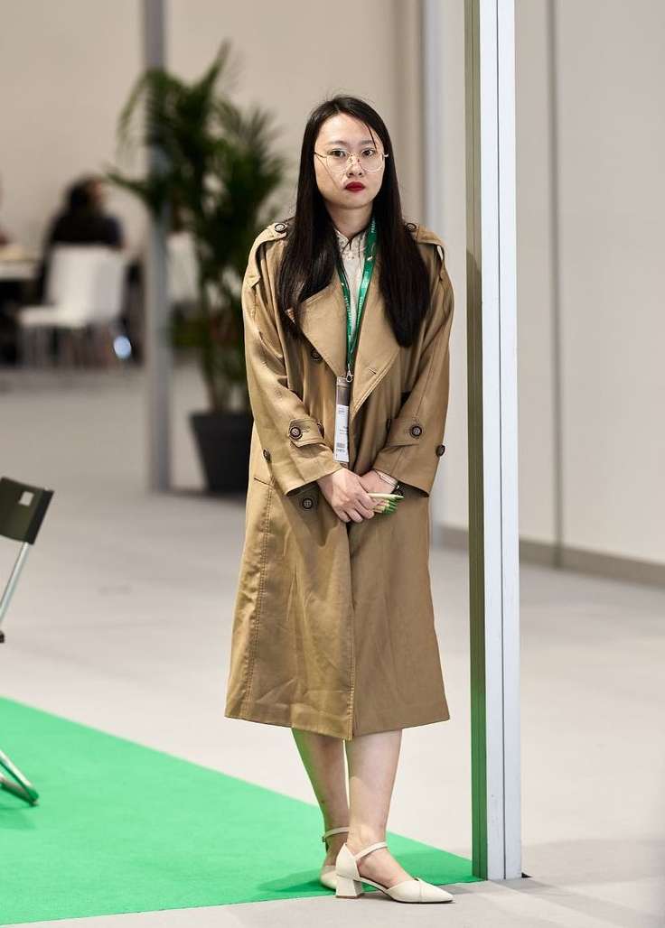 Adult woman in a tan trench coat stands in an indoor space with a neutral color palette, creating a contemporary and professional aesthetic.