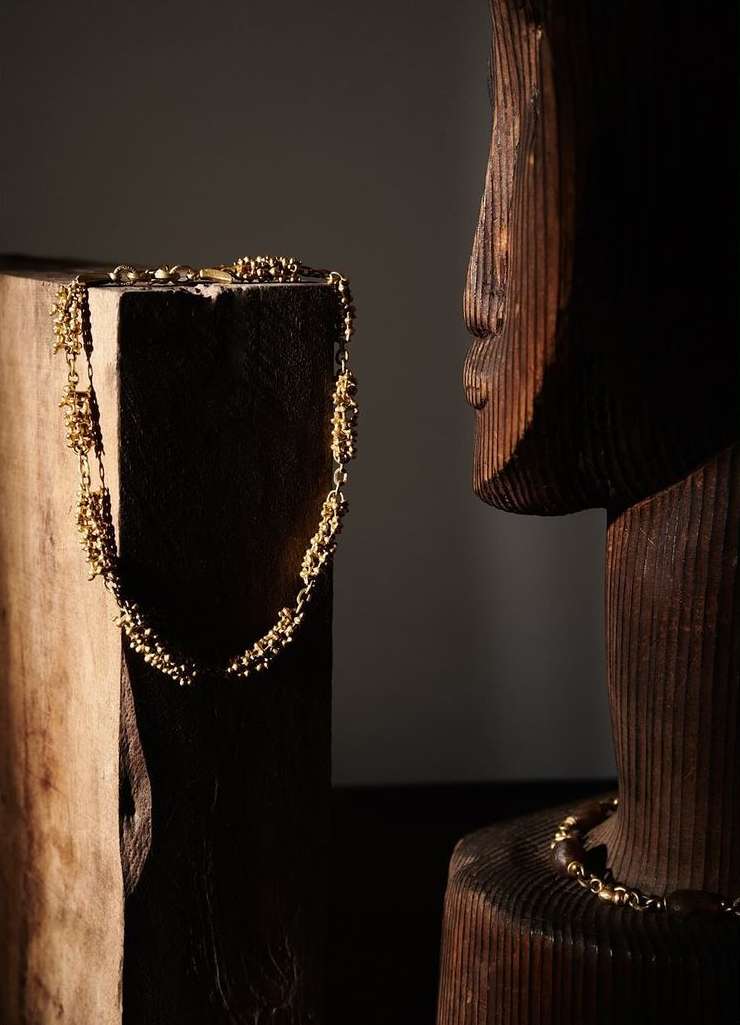 Golden beaded necklace displayed on a wooden block beside a carved wooden face in a softly lit, neutral-toned setting.