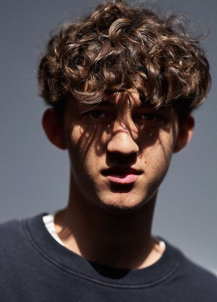 Close-up portrait of a young adult with curly hair wearing a black sweatshirt against a grey background in bright studio lighting.