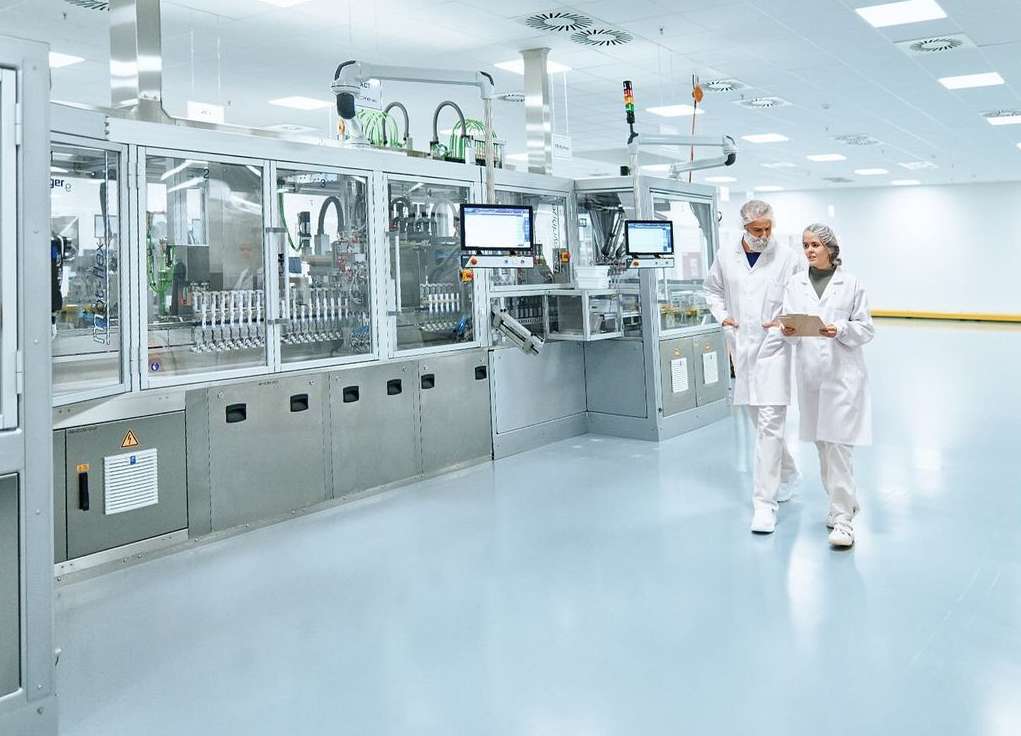 Two healthcare workers in lab coats walk past pharmaceutical manufacturing equipment inside a bright, sterile cleanroom environment.